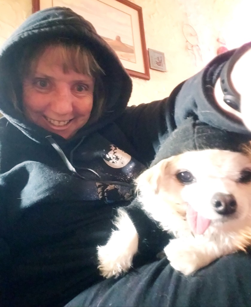 A smiling person wearing a black hoodie poses for a selfie with a small dog, also dressed in a matching black hoodie, on their lap.