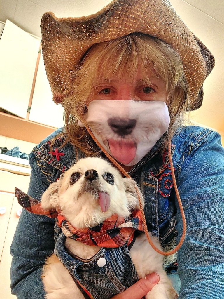 A smiling woman wearing a cowboy hat and a face mask with a dog's mouth design, holding a small dog that is also sticking its tongue out. They are in a cozy indoor setting.