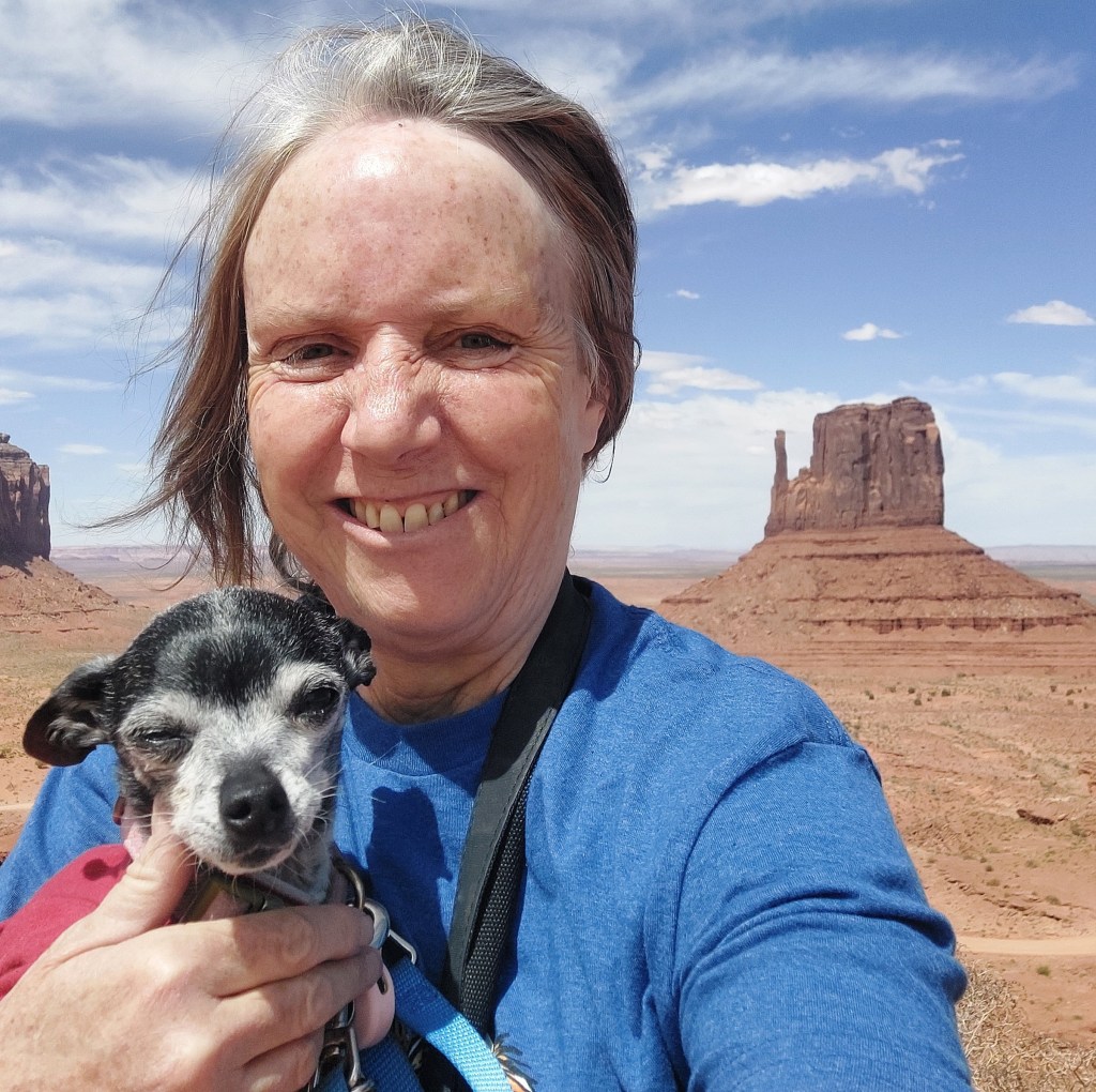 A woman with gray hair smiles while holding a small black and white dog, with a large rock formation in the background under a blue sky.