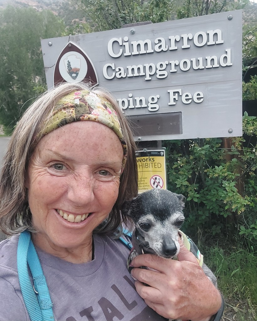 Smiling woman with a pet dog in front of the Cimarron Campground sign.