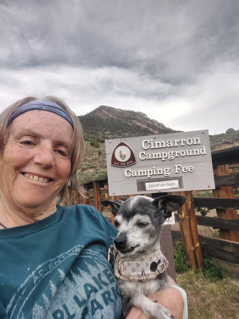 A woman smiling while holding a small dog, standing in front of a sign for Cimarron Campground, indicating a camping fee.