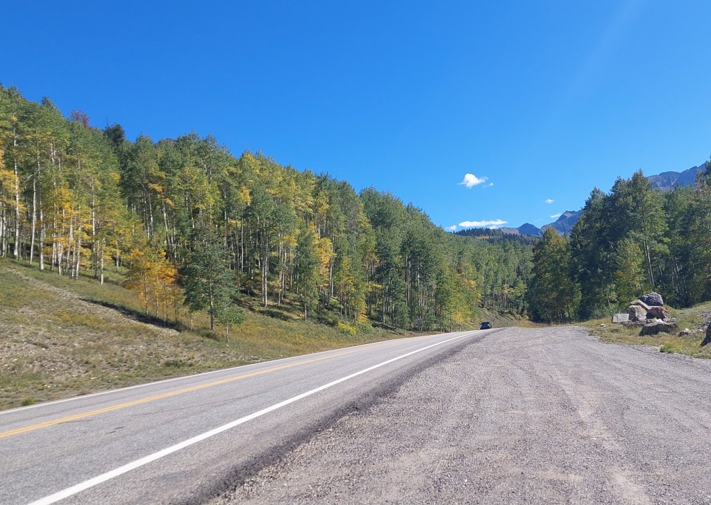 A wide view of a rural road surrounded by a colorful tree line in autumn, with blue skies and distant mountains.