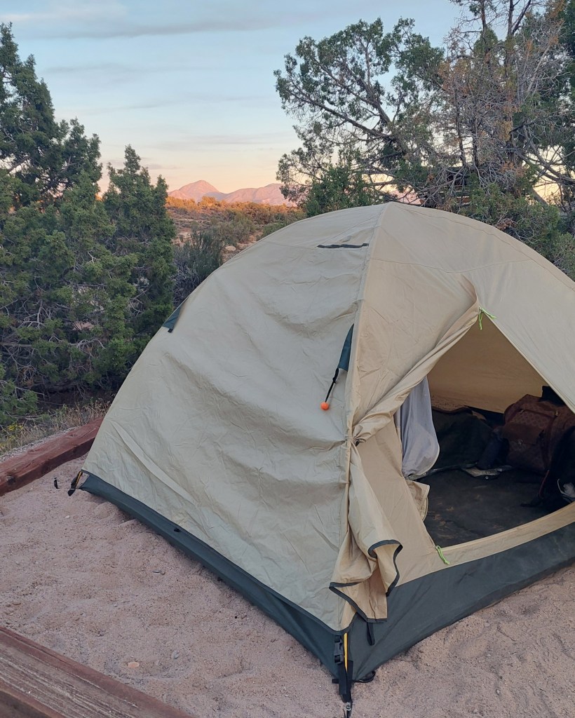 A camping tent set up on sandy ground, surrounded by trees and mountains in the distance under a pastel sky.