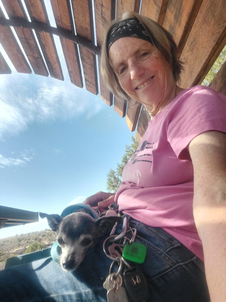 A woman with long hair and a headband smiles while sitting under a wooden structure, holding a small dog on her lap amidst a scenic outdoor background.