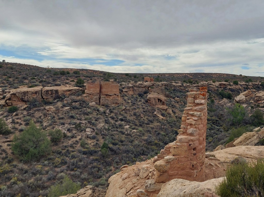 A rugged landscape featuring remnants of stone structures on a hillside, surrounded by desert vegetation and rocky terrain under a cloudy sky.