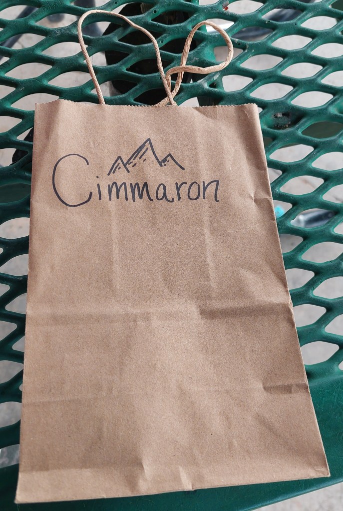 A brown kraft paper bag with the word 'Cimmaron' written in cursive and a mountain illustration, resting on a green metal table.