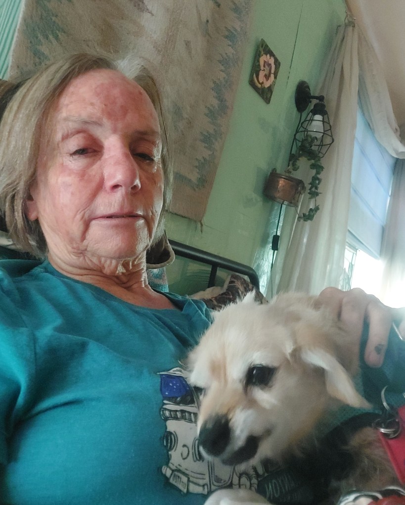 An elderly woman sitting on a bed, wearing a turquoise shirt, holding a small dog close to her chest with a gentle expression.