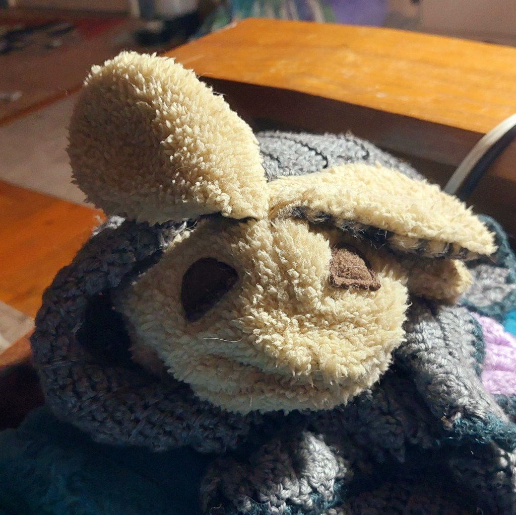 A close-up image of a soft, plush toy bunny head, partially covered with a crocheted blanket.